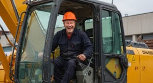 A construction worker wearing an orange hard hat and a dark work uniform is sitting inside the cab of a yellow excavator, laughing joyfully. He is gripping the control levers, appearing enthusiastic and engaged in his work. The background features industrial buildings and construction materials, indicating an active worksite environment. The worker’s expression conveys a sense of satisfaction and enjoyment in operating heavy machinery.