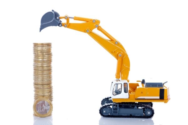 A miniature excavator toy positioned next to a tall stack of euro coins, symbolizing construction costs and financial investment in building projects.