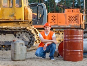 A construction worker in an orange safety vest and hard hat crouching beside fuel containers and an excavator at a construction site, smiling at the camera.