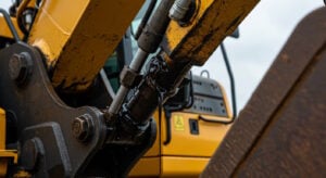 A close-up of a hydraulic system on a yellow excavator, showing a significant hydraulic oil leak from a damaged or loose hydraulic fitting. The black oil is visibly dripping down the metal components, covering the hydraulic cylinder and hoses. The background features the excavator’s cabin, with warning labels indicating maintenance concerns.