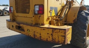 Close-up of the rear section of a yellow heavy construction vehicle showing rust, chipped paint, hydraulic components, and warning signs, indicating wear and maintenance needs.