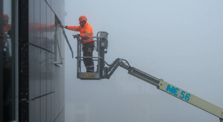 A construction worker in high-visibility orange safety gear and a hard hat uses a boom lift to perform maintenance on a building facade in dense fog, emphasizing the need for safety in low-visibility conditions.