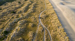 A drone captures a stunning aerial shot of a 4x4 vehicle driving along a winding dirt track through rolling coastal sand dunes. The golden grasses contrast with the soft, untouched sand on the right, creating a striking natural landscape. The remote and rugged terrain highlights the sense of adventure and exploration.