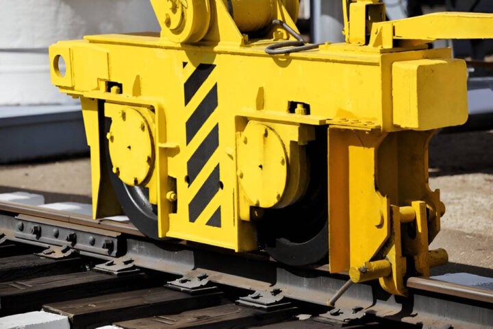 Detailed view of a yellow gantry crane wheel assembly mounted on steel rails. The assembly features a robust wheel system, safety markings with black diagonal stripes, and mechanical components used for guiding and supporting crane movement along a track.