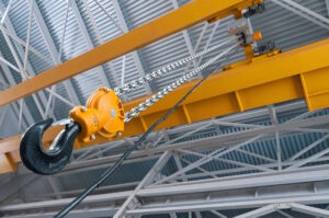 Detailed view of a heavy-duty overhead crane hook suspended by a strong metal chain inside an industrial facility. The yellow crane structure and steel roof beams are visible, highlighting the robust engineering of the lifting mechanism.