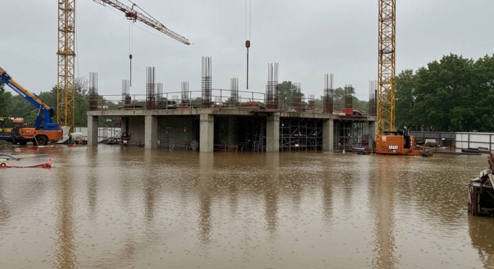 Flooded construction site with partially built concrete structure, cranes, and equipment surrounded by water on a rainy day.
