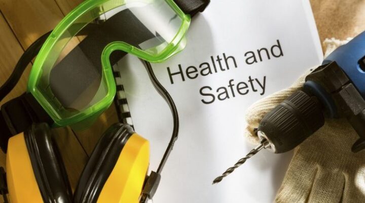 A set of personal protective equipment including green safety goggles, yellow earmuffs, gloves, and a power drill placed around a document labeled “Health and Safety” on a wooden surface, symbolizing workplace safety preparedness.