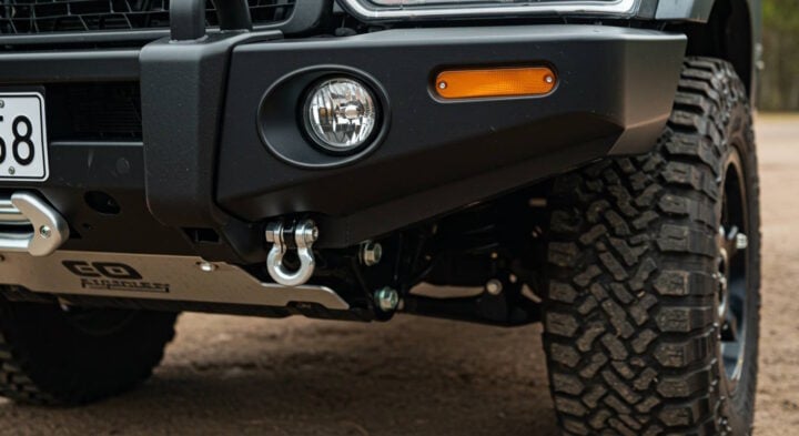 A close-up of the front bumper of an off-road vehicle, featuring a heavy-duty steel bumper with a built-in fog light, tow hook, and rugged all-terrain tires. The setup is designed for extreme off-road conditions.
