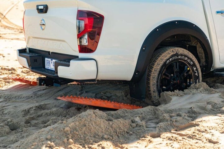 A white off-road pickup truck is stuck in deep sand, with traction boards placed under its rear wheels to aid in recovery. The vehicle’s tires are partially buried, and the sand shows signs of recent movement.