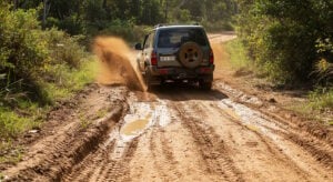A rugged off-road SUV drives down a muddy dirt road, kicking up a spray of dirt and water as it moves through a puddle. The vehicle is surrounded by lush green vegetation under bright sunlight.
