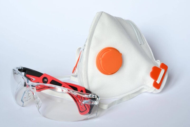 A white protective face mask with an orange exhalation valve and adjustable straps, placed next to a pair of transparent safety glasses with red and black frames. The personal protective equipment (PPE) is designed for respiratory and eye protection in hazardous environments.