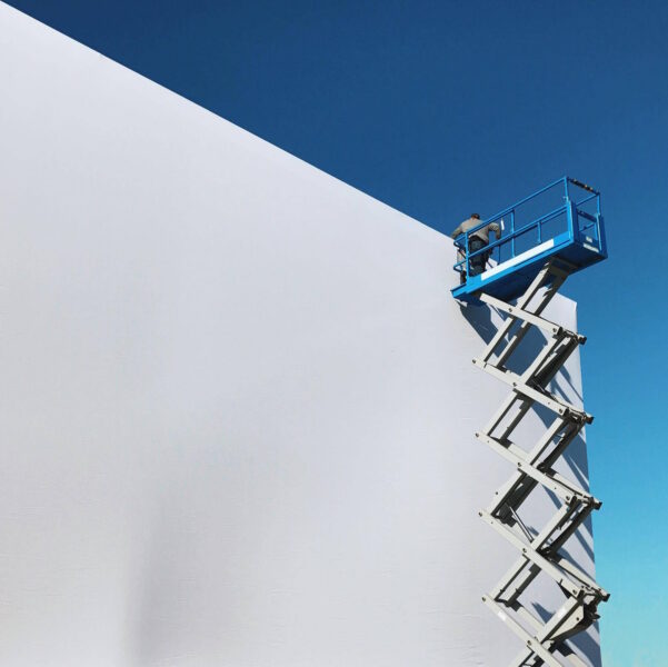 Worker on a blue scissor lift against a large white wall and clear blue sky