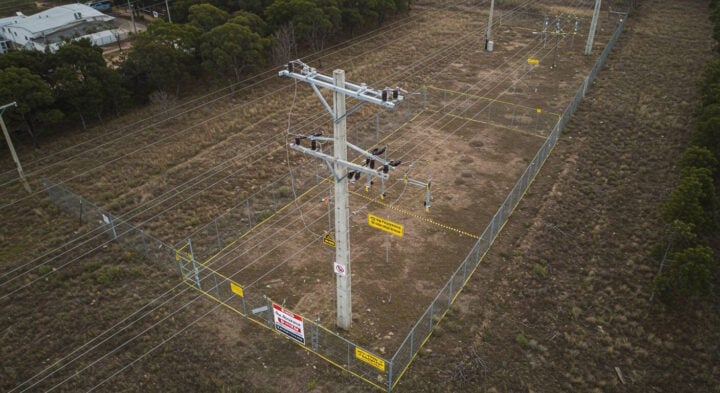 Aerial view of a fenced-off electrical substation with high-voltage power lines, surrounded by safety signage and clear barriers.