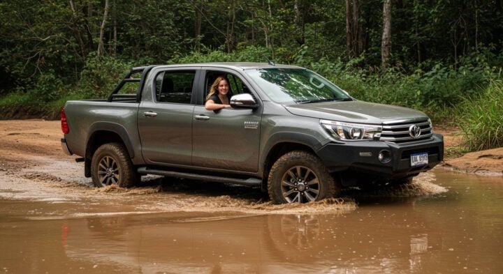 A woman with long blonde hair, wearing a black shirt, smiles from the driver's seat of a gray Toyota Hilux pickup truck as it drives through a muddy water crossing in a forested area. The truck's tires are partially submerged in the water, creating ripples as it moves forward.