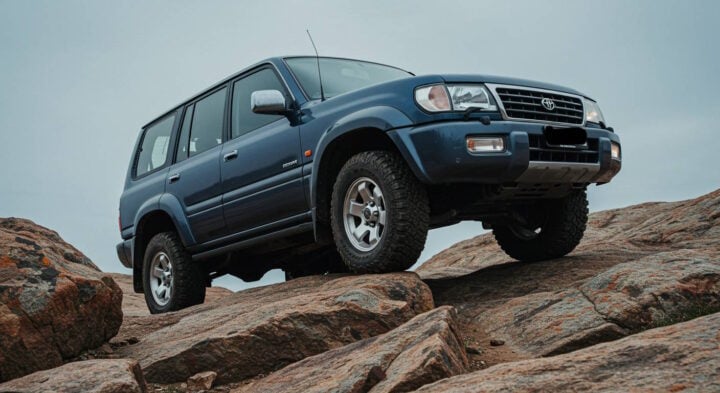 A blue Toyota Land Cruiser navigates a rugged rocky landscape, showcasing its off-road capability. The vehicle's lifted suspension and all-terrain tires emphasize its durability and readiness for extreme 4x4 adventures.