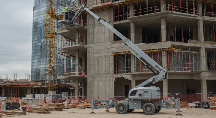 Construction worker on a Vertical Mast Lift at a high-rise building construction site with a modern glass skyscraper in the background.