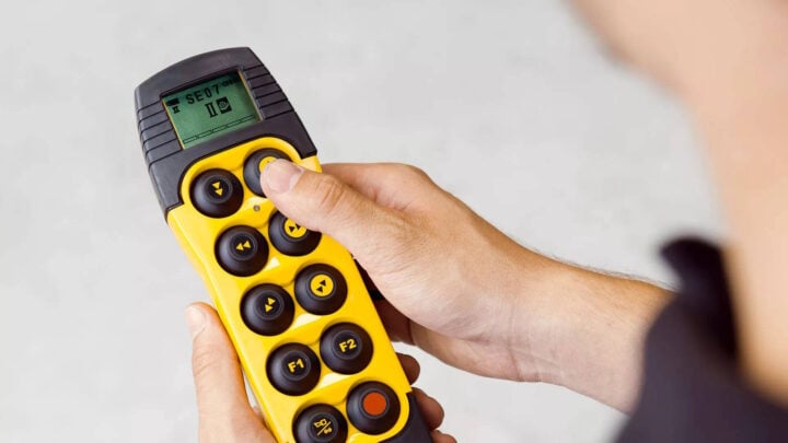 Close-up view of a person's hands operating a modern yellow and black wireless remote control unit for an industrial crane. The device features multiple directional and function buttons, along with a small digital screen displaying settings, illustrating the precision and safety offered by computerized crane systems.